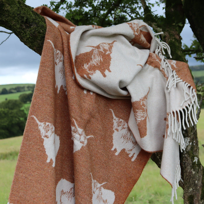Orange and beige reversible lambswool throw with printed Highland Cows draped over a tree