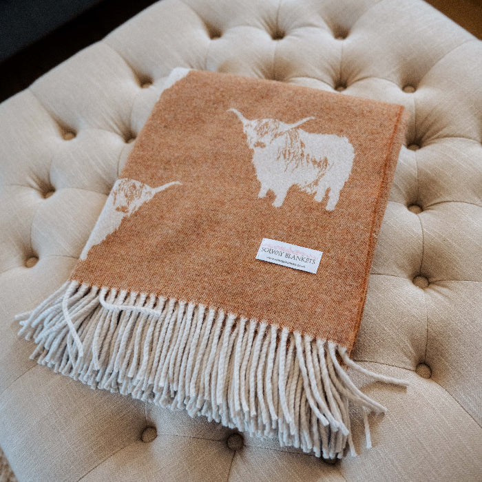 Orange lambswool throw with a Highland Cow design and fringe on a beige ottoman