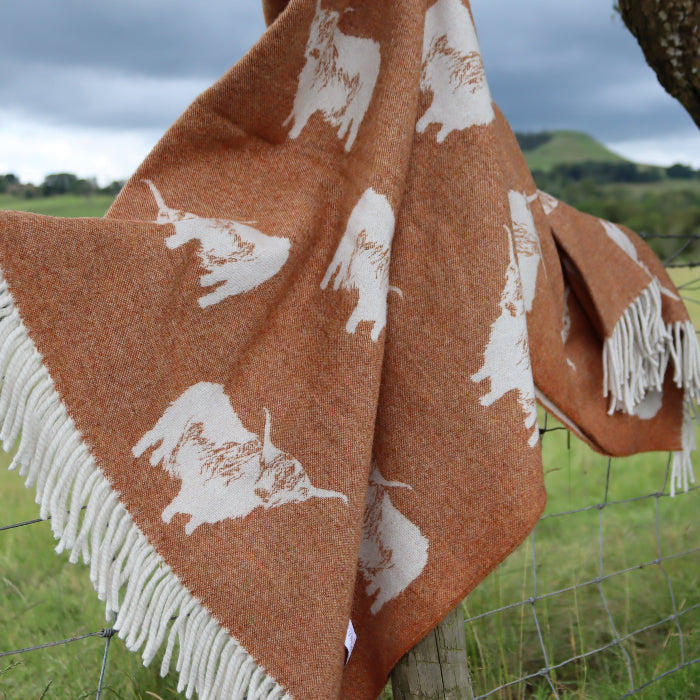Orange blanket with white Highland Cow patterns and fringe draped over a fence in a field