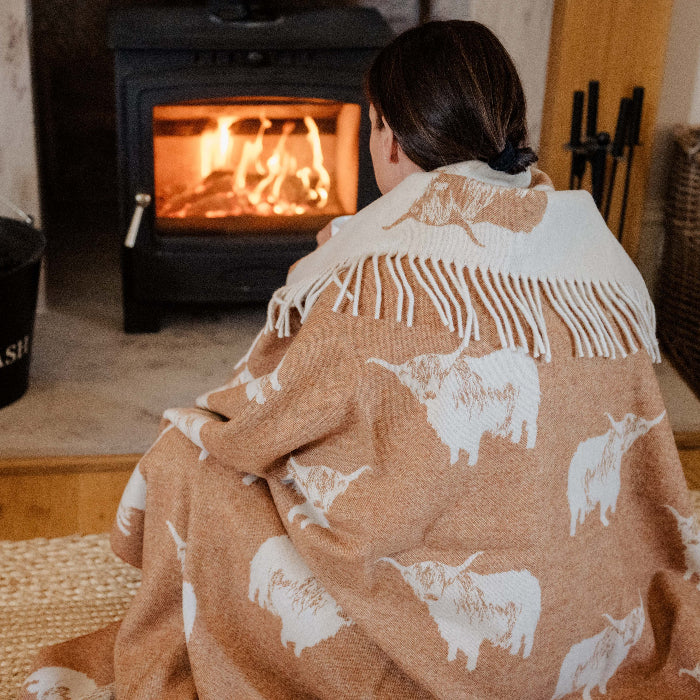 Person sitting infront of fireplace wrapped up in a cosy orange blanket with Highland Cow patterns