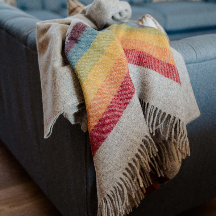 Highland Horizon Shetland Wool Rainbow Stripe Blanket colorful striped blanket draped over a gray sofa with a blurred cosy background