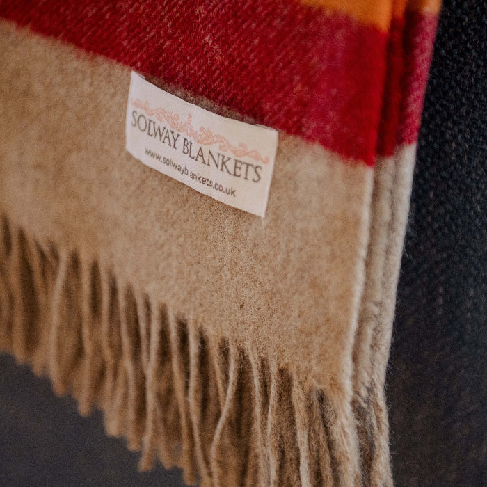 Close-up of Highland Sunrise Shetland Wool blanket showing part of rainbow stripe, fringes and a visible Solway Blankets label on a dark background