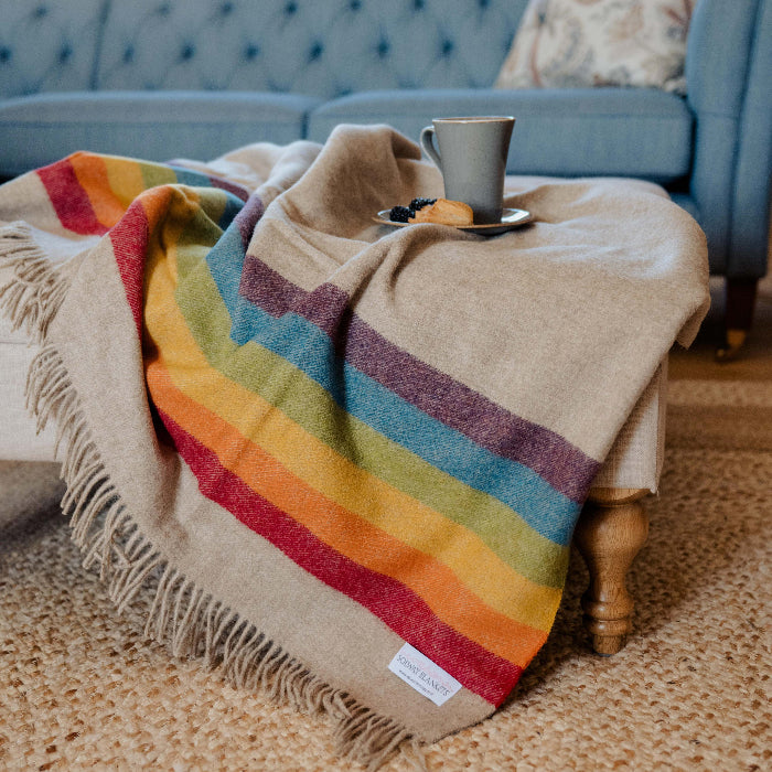 Highland Sunrise Beige Blanket with colourful rainbow stripe draped over a coffee table with a cup and cookies in a cosy living room.