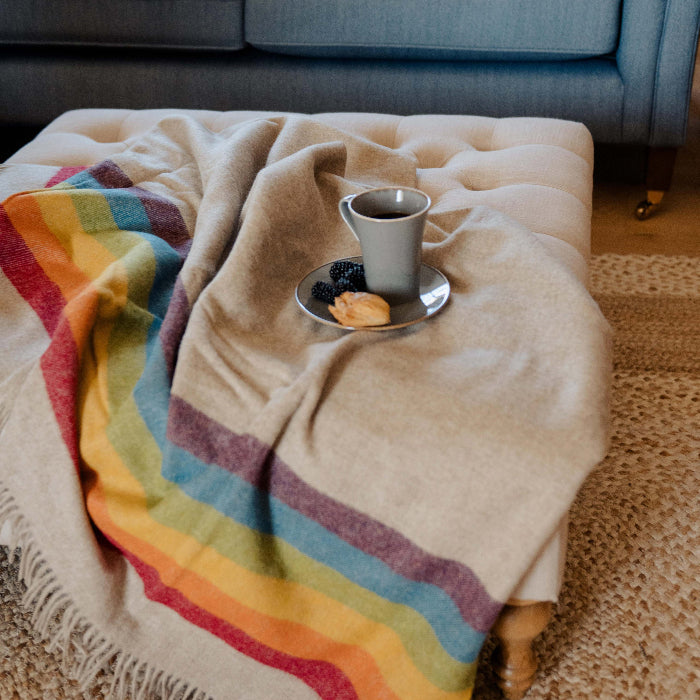 Colorful beige blanket with rainbow stripes draped over a ottoman with a cup and saucer on top, in front of a blue tufted sofa.