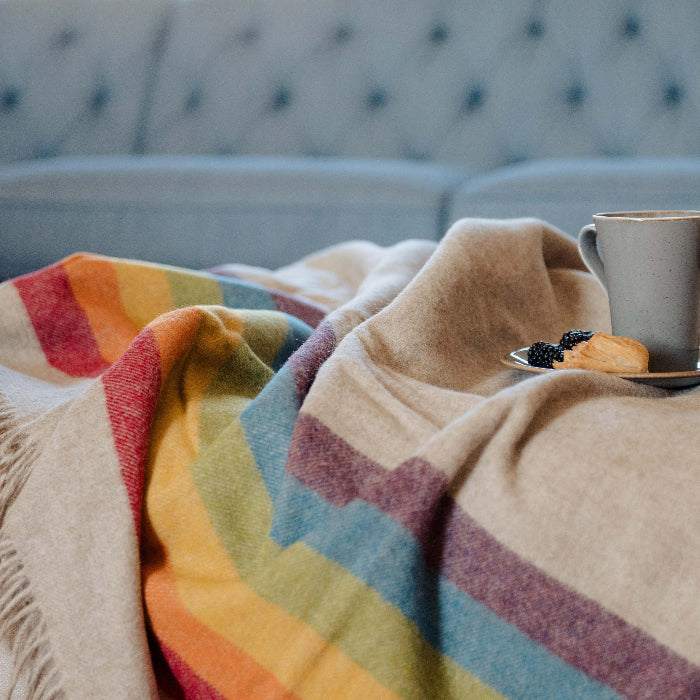 Highland Sunrise Beige Shetland wool blanket with rainbow stripe draped over a surface with a cup and plate on top, in front of a blue upholstered chair.