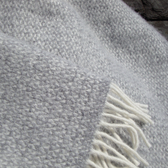 Close up of a grey textured fabric wool blanket with white fringe against a stone background