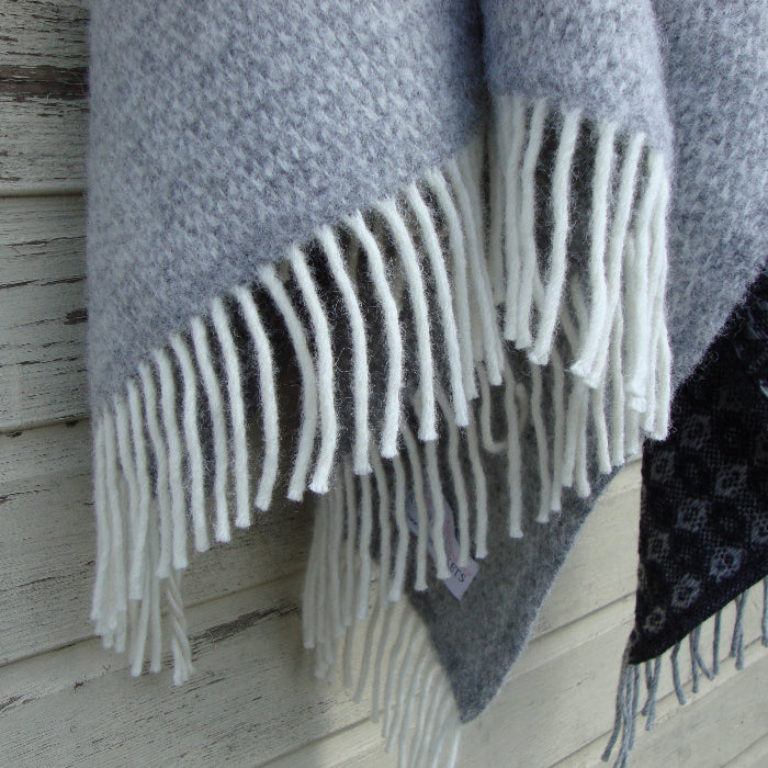 Close up of a grey wool blanket with white fringe draped over a wooden surface