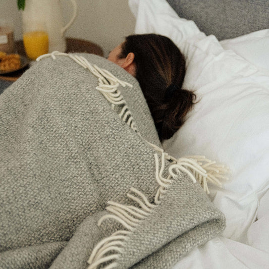 Person lying in a grey bed with white furnishings wrapped up in a grey wool blanket with white fringe with a side table of pastries and orange juice