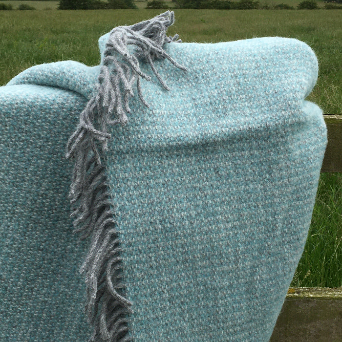 Spearmint and grey soft wool blanket throw with frayed fringes draped over a wooden fence with a natural background