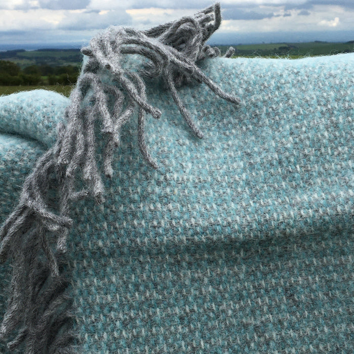 Close up of a spearmint and grey soft wool throw with frayed fringe with a grassy field in the background