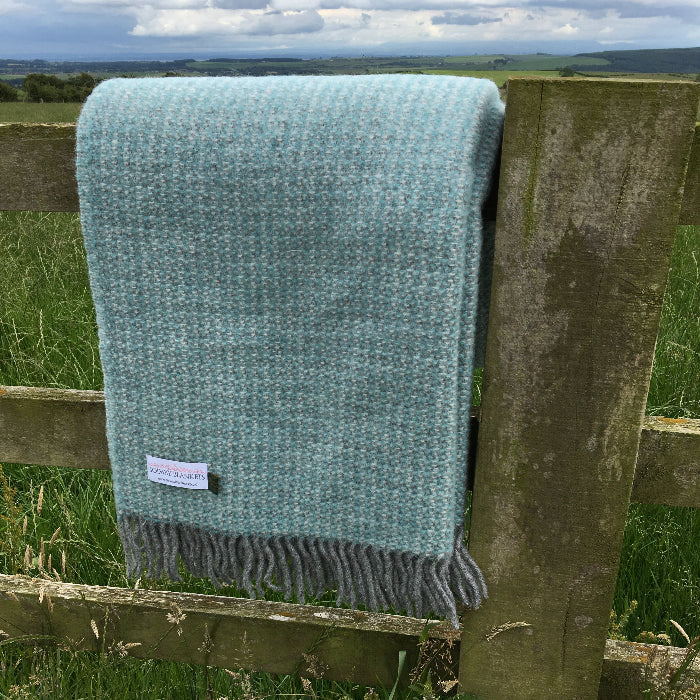 Spearmint and grey soft woven wool blanket folded over wooden fence set in a grassy field