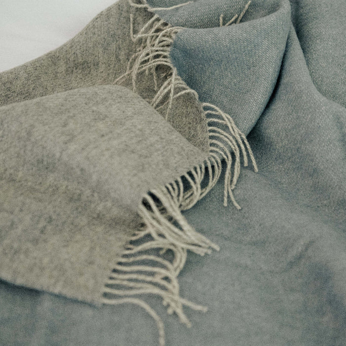 Blue and light grey reversable wool blanket with fringes loosely placed on white bedding