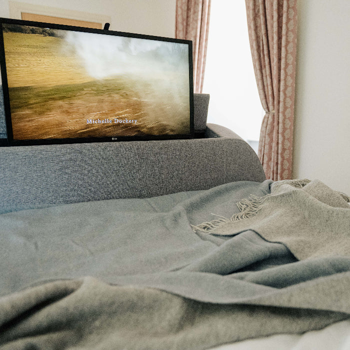 Blue wool throw with a light grey on the reverse, draped over a bed with a television displaying a nature scene in the background.
