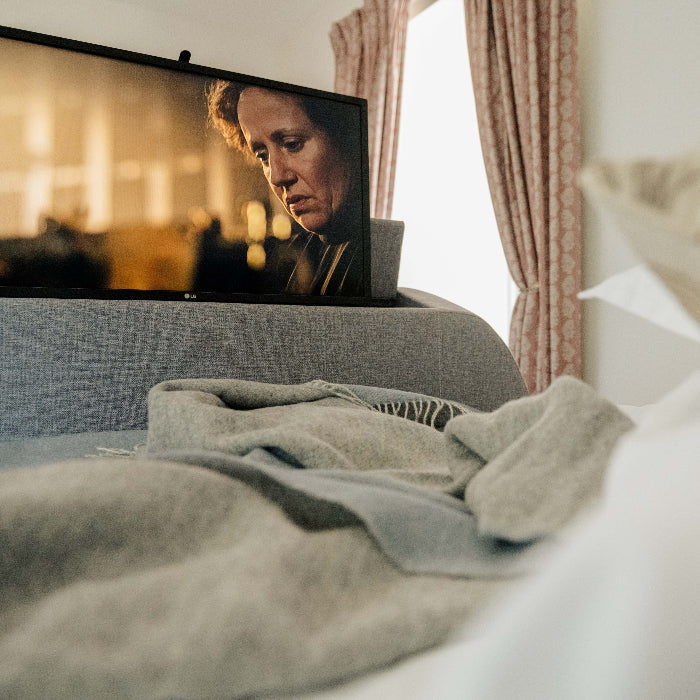 Program on the television in a bedroom with a blue and light grey reversable blanket and decorative cushion. 