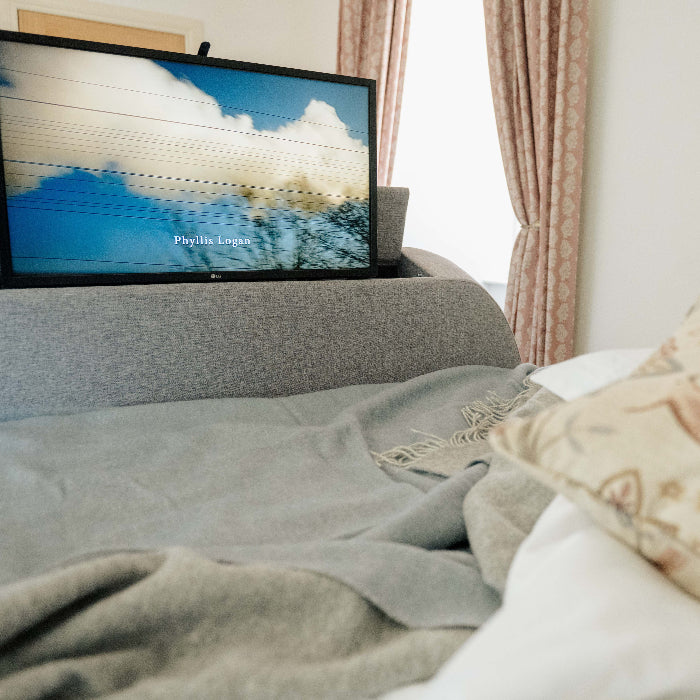 Grey bed with a television and a decorative pillow, featuring a blue wool throw with a soft grey colour on the reverse lying on the bed.  