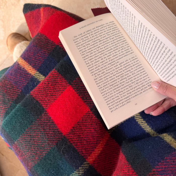 A person holding an open book with the Kilgour Tartan Knee Rug in red, green, and blue laid over the knee. A hand with painted nails is visible holding the book.
