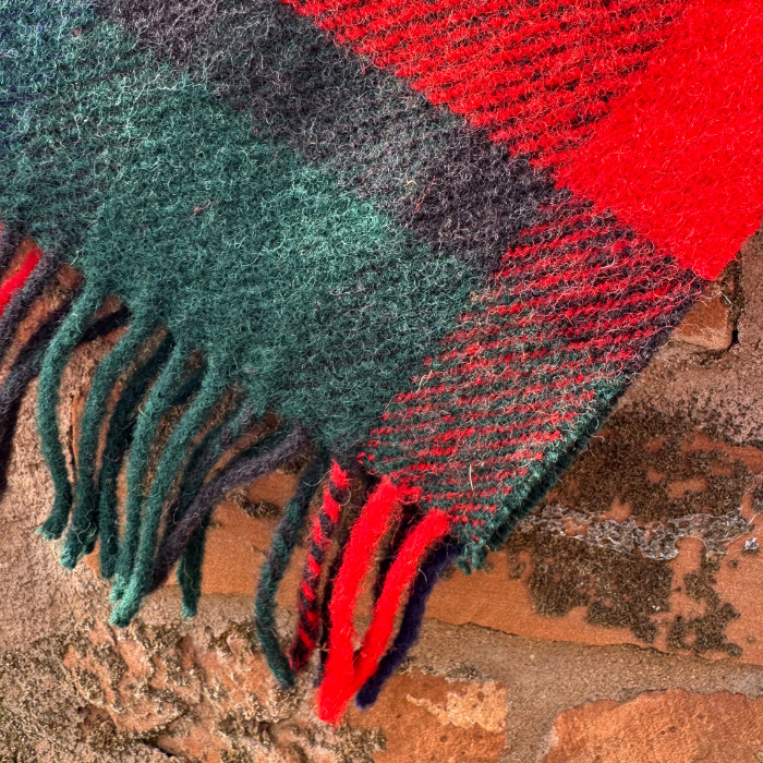 A close-up of a Kilgour tartan knee blanket with fringe, featuring red, dark green, and black stripes, draped over a rustic brick surface.