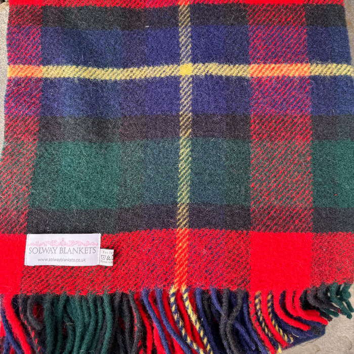 A close-up of a Kilgour Pure Wool Knee blanket with fringe, featuring red, green, navy, black, and yellow Tartan. A white woven label reading 'SOLWAY BLANKETS' is visible near the bottom left.