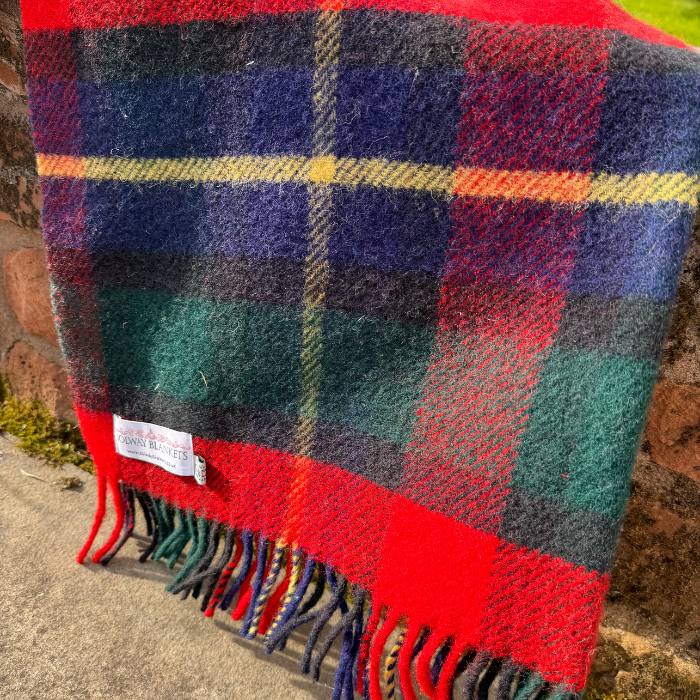 A close-up of a woven Kilgour Tartan blanket draped over a stone wall, showing a multicolor tartan pattern with red, navy, green, black, and yellow stripes, finished with fringed edges and a visible Solway Blankets brand label.