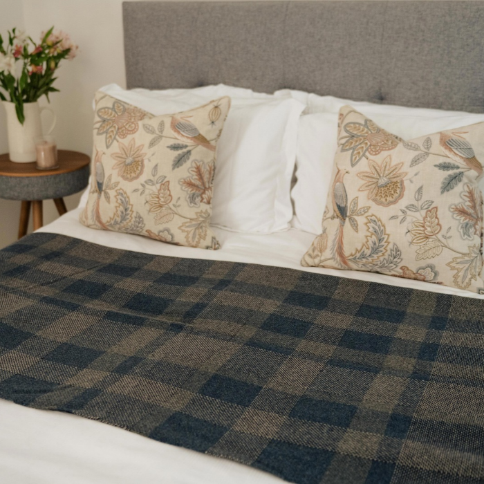 Lochmaben navy and grey checked lambswool blanket is spread across a bed with white sheets, flanked by two beige floral throw pillows against a grey upholstered headboard.