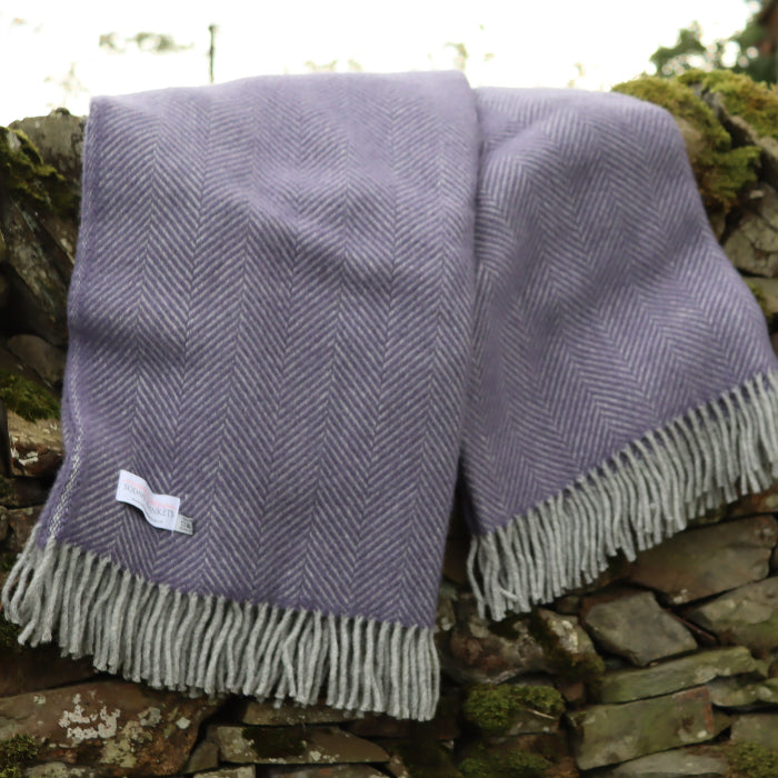 Lilac and silver herringbone style wool blanket with silver fringe, draped over a stone wall. 