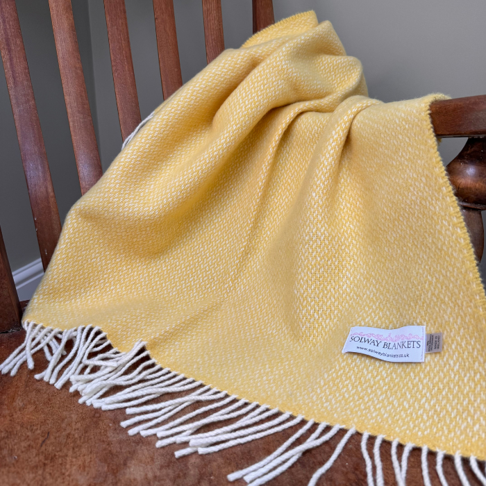 A soft pale yellow woven cashmere baby blanket with white fringe, draped over a wooden chair. A small woven label on the blanket reads 'SOLWAY BLANKETS'.
