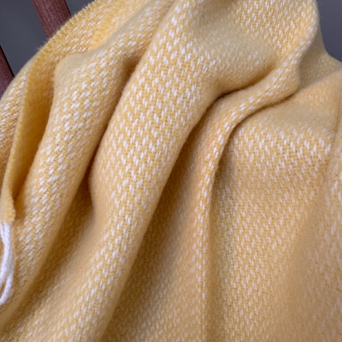 A close-up of a soft woven cashmere and merino soft baby blanket in a pale buttery yellow with a subtle white textured pattern, draped in folds.