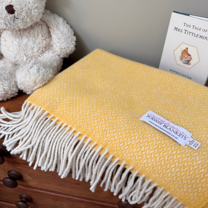 A yellow and white textured woven throw blanket, in super soft cashmere and merino wool with white fringe, displayed on a wooden surface next to a white teddy bear and a book.