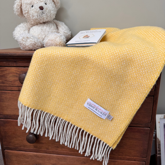 A soft yellow woven cashmere mix baby blanket with white fringe is draped over a dark wooden dresser. A cream teddy bear sits on top of the dresser beside a small book, and a visible fabric label reads 'Solway Blankets'