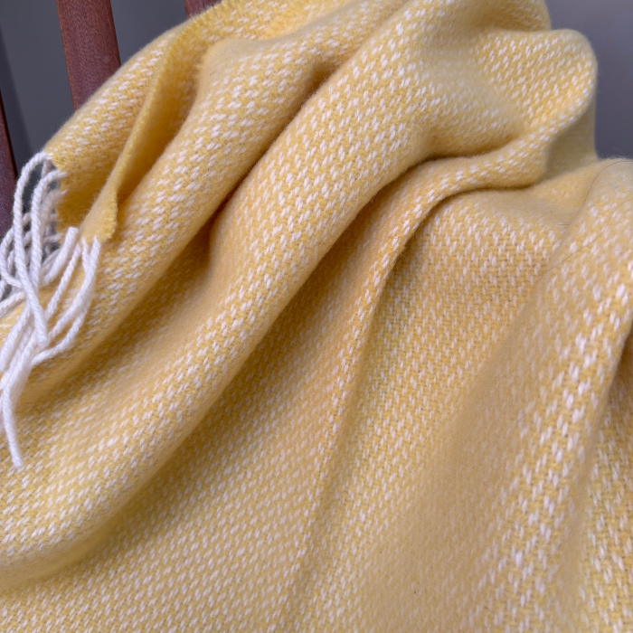A close-up of the textured weave of a soft cashmere mix baby blanket in pale yellow and white with white fringe tassels, draped in folds.