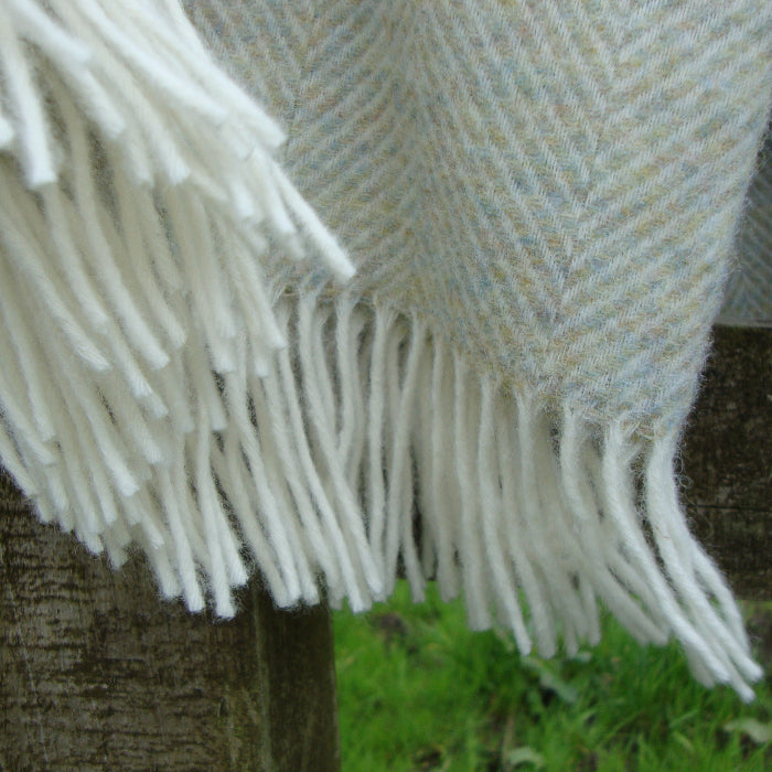 Textured Light Sage Green Herringbone Shetland Wool Blanket with white fringe draped over a wooden surface