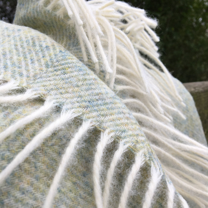 Close-up of a textured Light Sage Green Herringbone Shetland Wool Blanket with white and mottled green weave, outdoor setting.