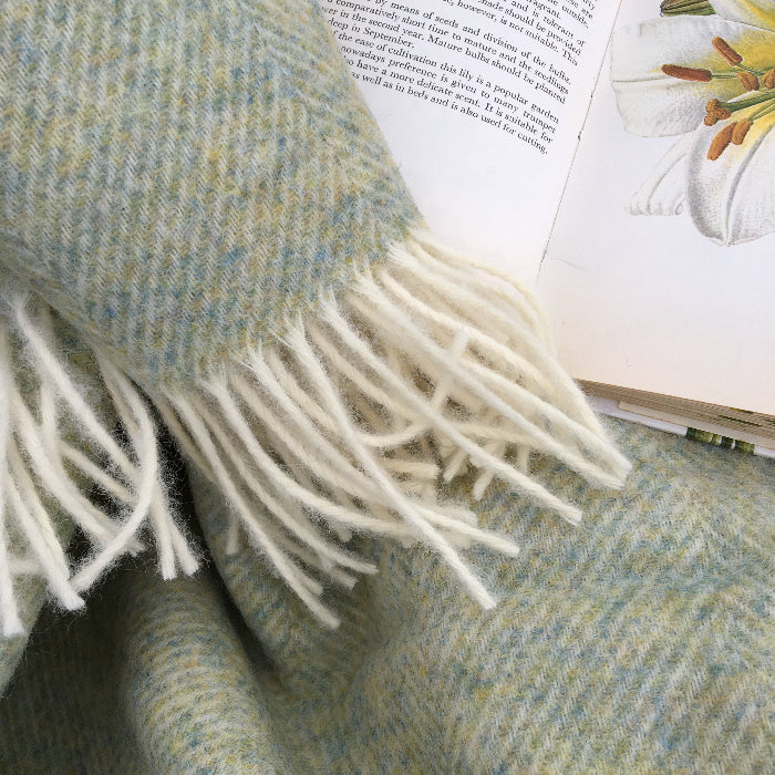 Light Sage Green Herringbone Shetland Wool Blanket a British woven blanket with fringes on a surface next to an open book.