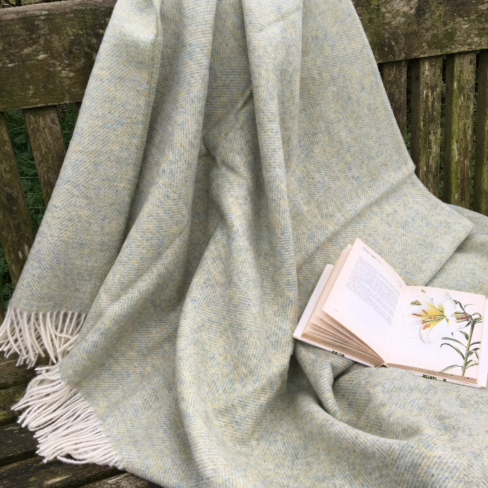 Light Sage Green Herringbone Shetland Wool Blanket a Green woven blanket draped over a wooden surface with a book on top
