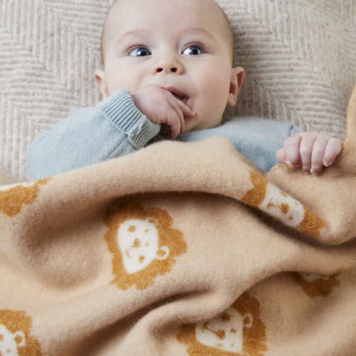 Baby lying on the Lion Caramel Merino Wool Baby Blanket a beige blanket with lion patterns, wearing a light blue sweater.