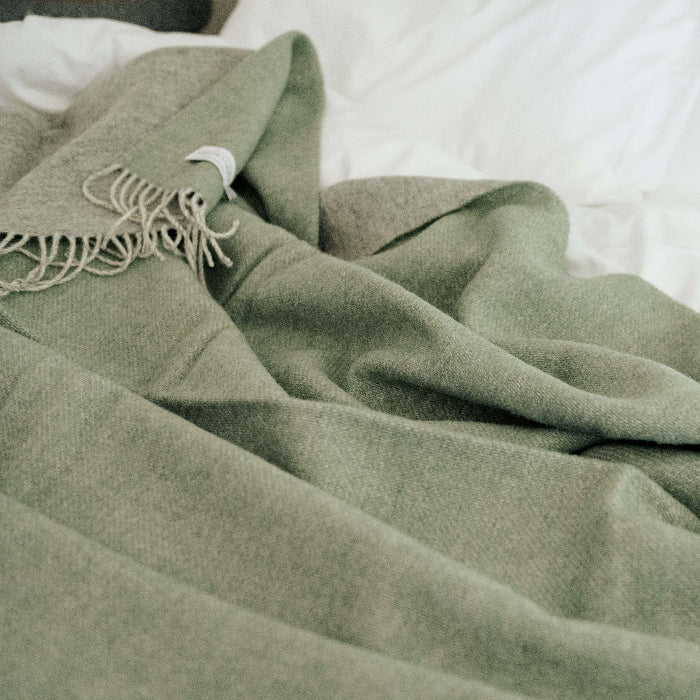 Green wool throw with a soft grey on the reverse, draped over a white bed