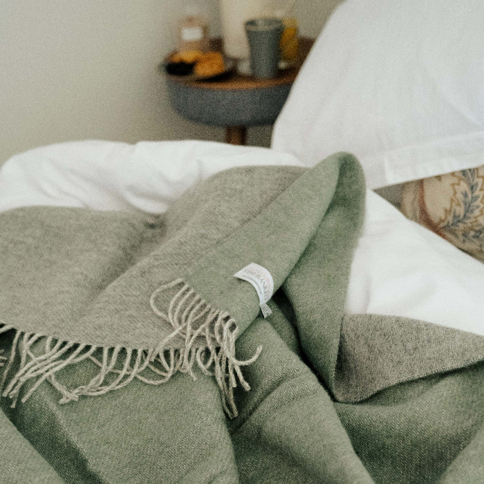 Green blanket with a soft grey reverse with fringes draped over white bedding, with a blurred background of a bedroom.