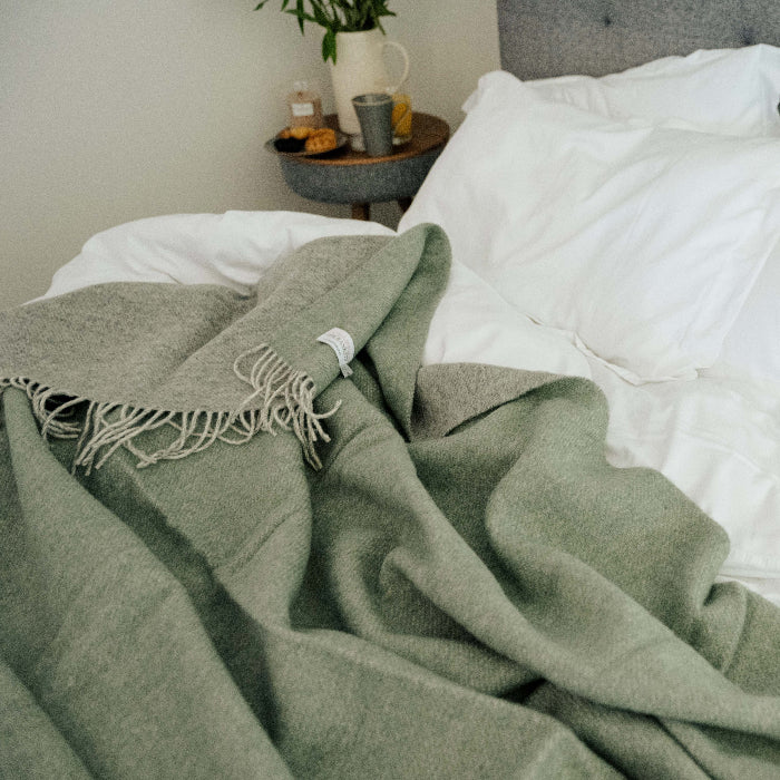 Green wool throw with a soft grey colour on the reverse, draped over a bed with white pillows and a gray headboard.