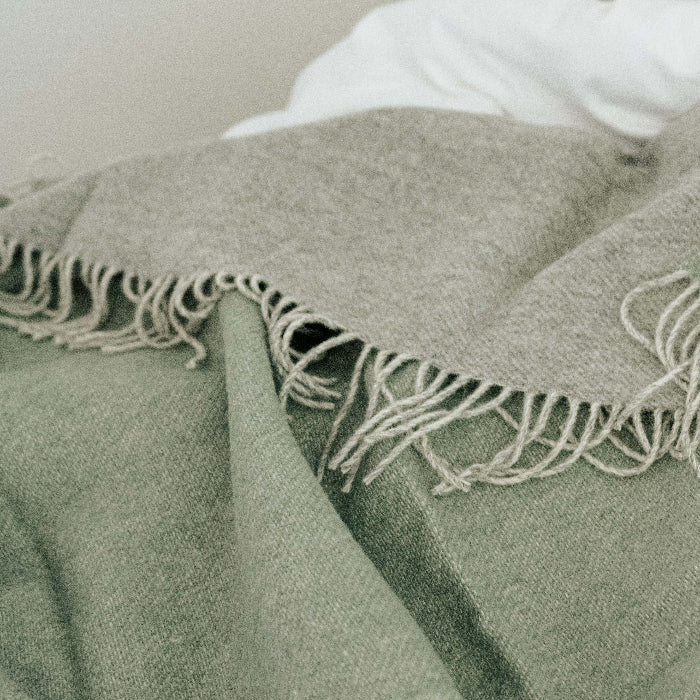 Green wool blanket with a soft grey colour of the reverse with fringe placed on a white bed 