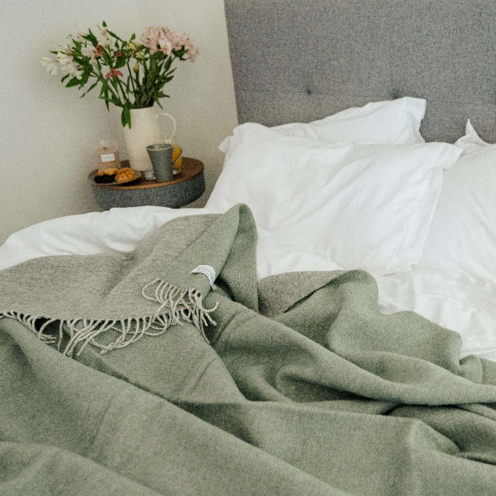 Grey bed with white bedding and a green throw blanket with a light grey colour on the reverse, featuring nightstand with flowers in the background