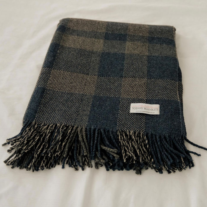 A folded Lochmaben Scottish Woven Lambswool  blanket with fringe ends, laid flat on a white fabric background. The blanket features a checkered pattern in dark navy blue, charcoal grey and off white, with a small woven label reading 'Solway Blankets.