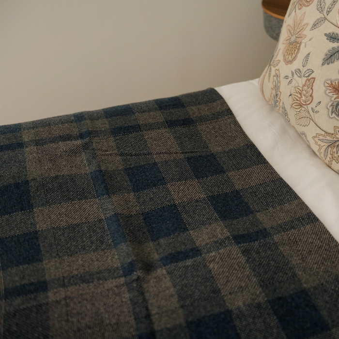 Close-up of the textured lambswool weave and classic check pattern of the Lochmaben throw, draped over a bed, with a white sheet and a patterned cream pillow visible.