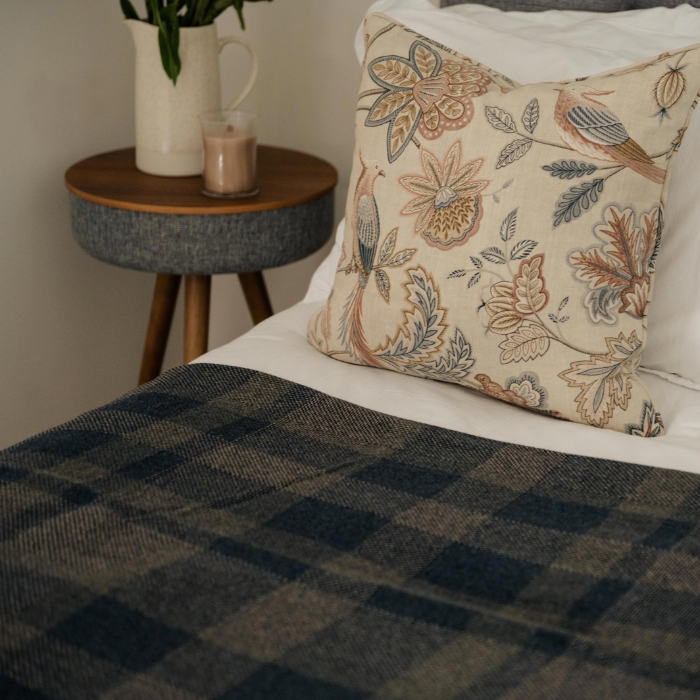 A styled bedroom scene showing the Lochmaben navy and grey checked lambswool throw spread across a bed, with a cream pillow featuring a botanical and bird pattern leaning against the headboard. A small wooden side table with a vase and candle sits beside the bed.