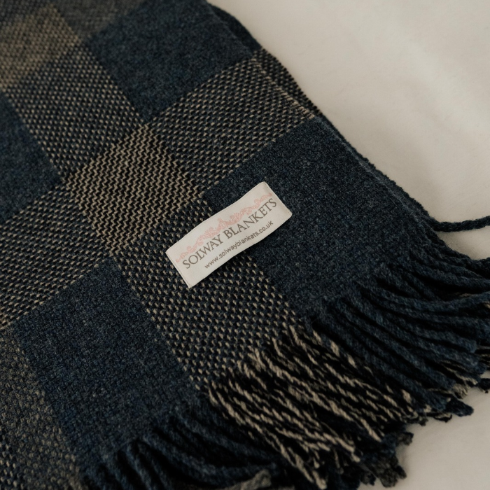 A close-up of the Lochmaben Scottish lambswool throw in navy, blue-grey and charcoal check with twisted fringed edges, featuring a white woven label that reads 'SOLWAY BLANKETS'.