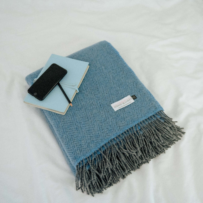 Folded blue cashmere throw with fringes on a white surface, featuring a phone and notebook and Solway Blankets label.