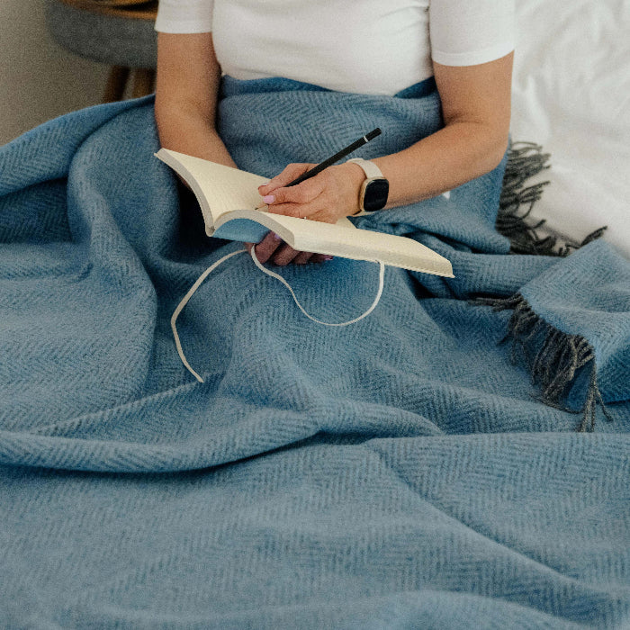 Person sitting on a bed covered with a blue cashmere throw, holding a notebook and pencil.