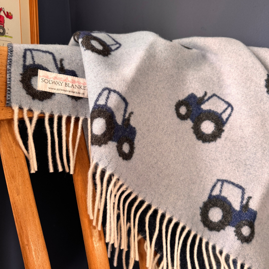 A light blue woven merino baby blanket with cream fringe and a repeating tractor pattern in navy and blue, draped over a wooden chair. A woven label reads 'SOLWAY BLANKET'.