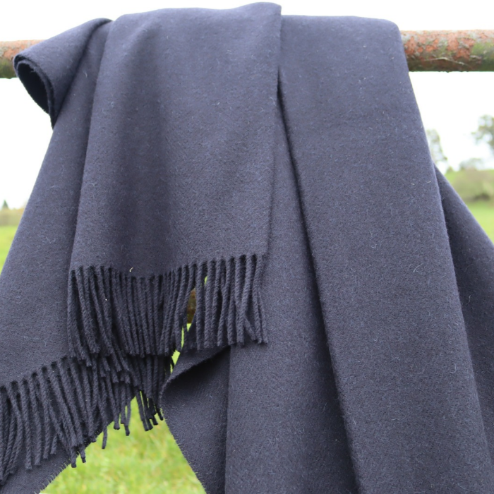 Moffat Midnight pure cashmere throw in deep navy blue with fine weave and fringed edge, draped over a rusty metal pole with a blurred green grassy field in the background.