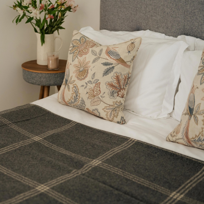 A styled bedroom scene showing a bed with white bedding, a soft blue grey cashmere throw woven and finished in Scotland with two decorative throw pillows featuring a botanical and bird pattern on a beige background. A small side table with a vase of flowers is visible in the background.