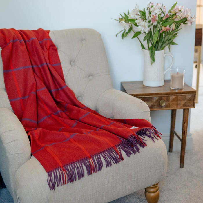 Luxury pure cashmere red throw with purple windowpane over check.  Woven in Scotland.  Beautifully styled, draped over a button backed, linen covered bedroom chair with a side table, vase of flowers and candle.